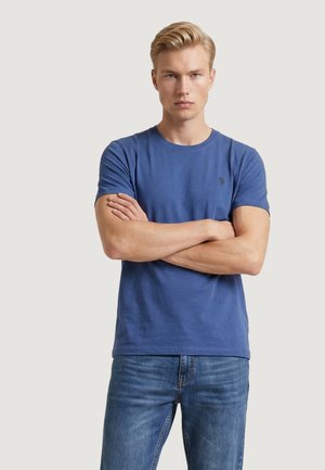 Giovane uomo con capelli biondi, che indossa una maglietta a maniche corte blu e jeans blu, in piedi con le braccia incrociate su uno sfondo uniforme.