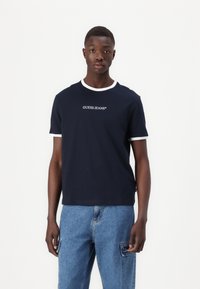 Camiseta de manga corta azul marino con ribete blanco, que presenta el logo "GUESS JEANS" en la parte frontal, fabricada en material de algodón, con un ajuste clásico.