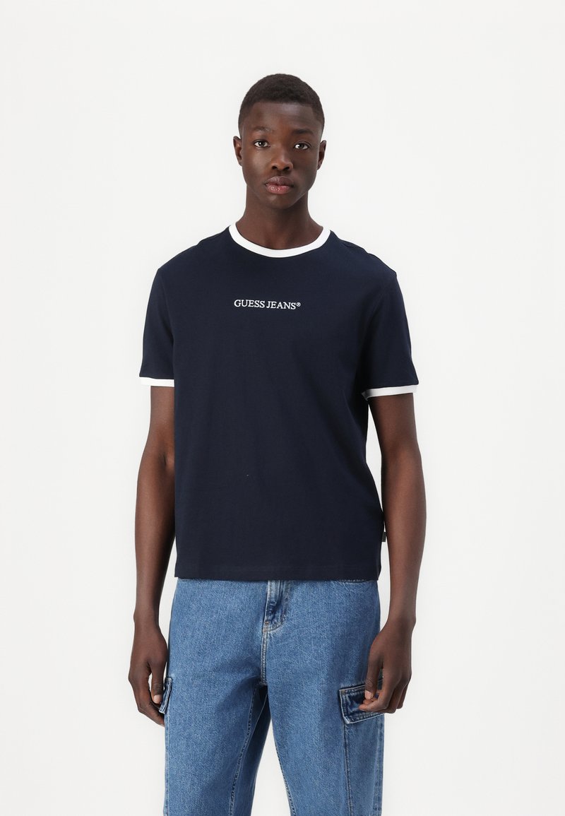 Camiseta de manga corta azul marino con ribete blanco, que presenta el logo "GUESS JEANS" en la parte frontal, fabricada en material de algodón, con un ajuste clásico.