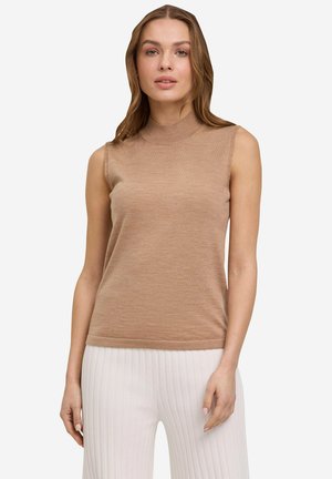 Donna che indossa un top marrone chiaro senza maniche con collo alto e pantaloni bianchi plissettati a gamba larga, in piedi su uno sfondo semplice.