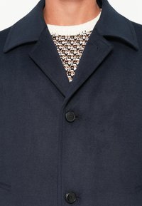Primo piano di un cappotto di lana blu navy con due bottoni neri indossato sopra un maglione lavorato a maglia multicolore con motivi bianchi, neri e marroni.