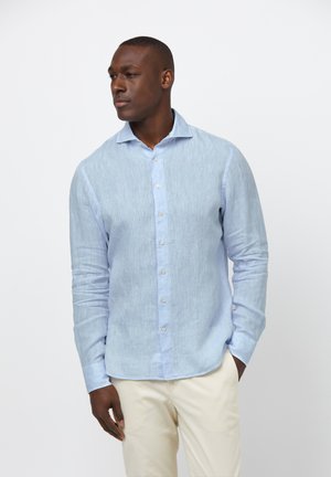 Zwarte man staat met handen in de zakken, draagt een lichtblauw overhemd met knopen en beige broek, kijkt opzij tegen een effen achtergrond.