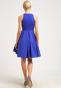 Modré šaty bez rukávů s přiléhavým živůtkem, rozšířenou sukní a plisovaným zadním detailem. Látka působí hladce s jemným leskem.