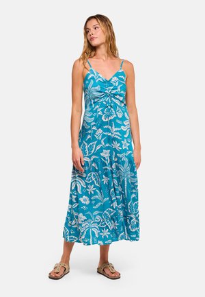 Mujer de pie, con un vestido azul sin mangas con estampado floral tropical blanco y sandalias color beige, mirando ligeramente hacia un lado.