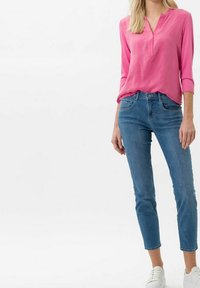 Blusa rosa a maniche lunghe con scollatura a V e polsini arrotolati, abbinata a jeans blu a gamba stretta e sneaker bianche su uno sfondo neutro.