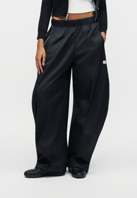 Fekete, széles szárú nadrág fényes anyagból, gumis derékrésszel és egy kis logófolttal a bal oldalon.