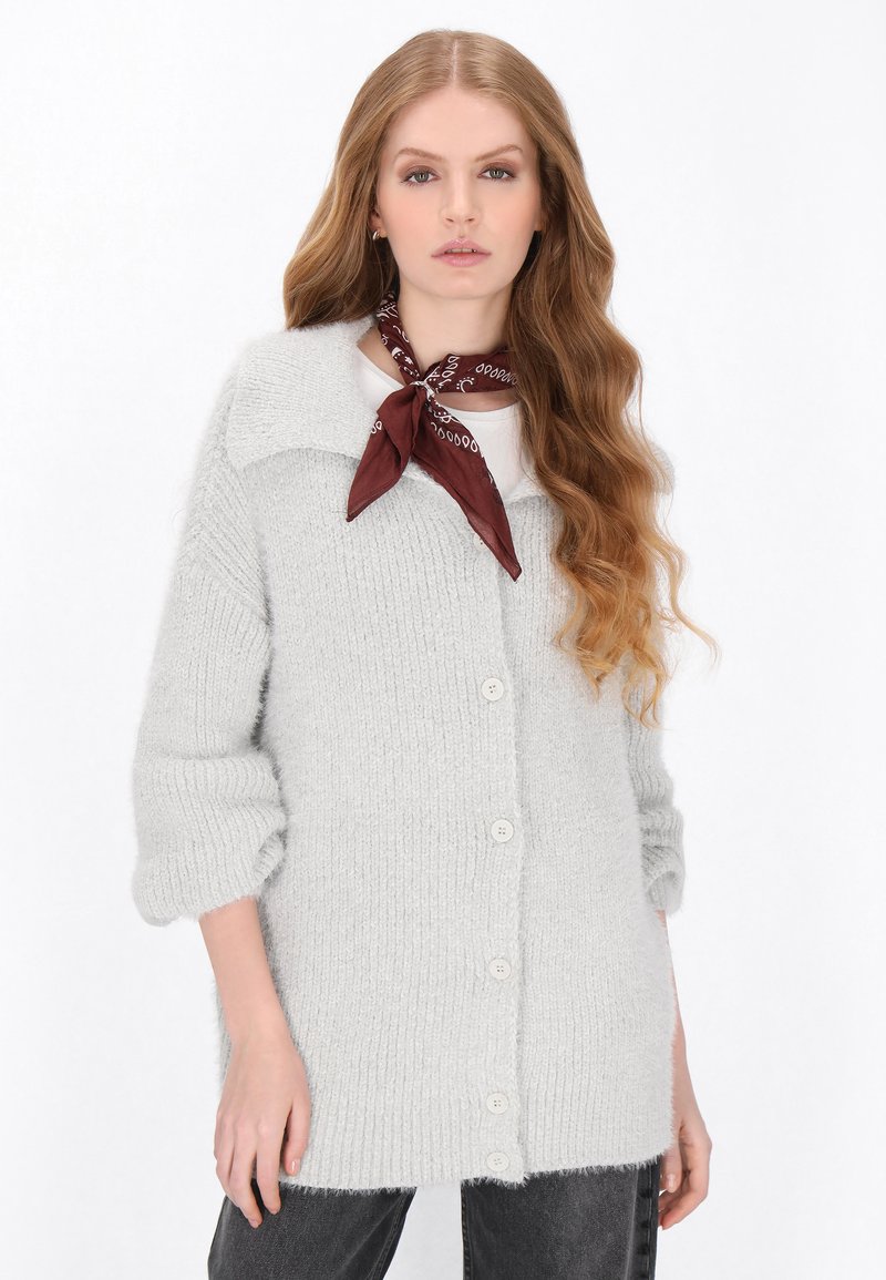 Šedý pletený cardigan s volným střihem, s texturovanou tkaninou, širokým límcem a pěti předními knoflíky. Nosený přes bílé tričko s hnědým šátkem.