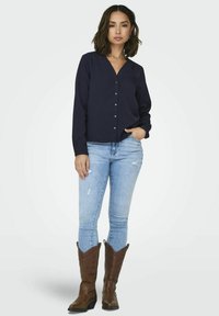 Chemisier bleu marine à boutons avec un col en V, manches longues, associé à un jean skinny bleu clair déchiré et des bottes de cowboy marron.