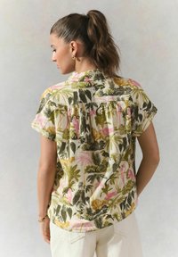 Korte mouwen, knoopshirt met een tropisch patroon van rozenflamingo's, groene palmbomen en zwarte vogels op een crèmekleurige achtergrond.