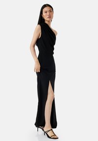 Schwarzes Kleid mit einem Schulterriemen, drapiertem Design, tailliertem Schnitt, Schlitz bis zum Oberschenkel und glatter Textur. Kombiniert mit schwarzen Riemchen-Heels.