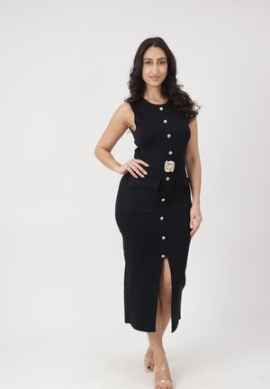 Frau mit langen dunklen Haaren, die ein ärmelloses schwarzes Midikleid mit goldenen Knöpfen und transparenten Riemchenabsätzen trägt, steht vor einem weißen Hintergrund.