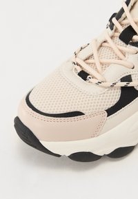 Sko med mesh-overdel i lys beige, med svarte og hvite detaljer, teksturerte overlegg og en tykk svart gummisåle.