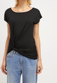 Žena oblečená v přiléhavém černém tričku s krátkým rukávem a uzlem na přední straně a světle modrých džínách s vysokým pasem, stojící před jednofonním pozadím.