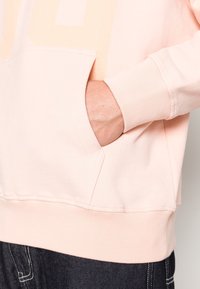 Jasnoróżowa bluza z kapturem z kieszenią kangurką, ściągaczami na mankietach i dole, o miękkiej fakturze i minimalistycznym designie. W zestawieniu z ciemnymi dżinsami.