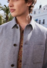 Grå, struktureret jakke med krave og to knapper, båret over en mønstret rød og brun skjorte. Har en brystlomme og en herringbone-designet.