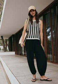 Woman wearing black and white sleeveless top, black loose pants, beige cap, flip-flops, and carrying a striped tote bag, standing on a patio.