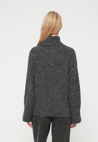 Suéter cinza escuro, de corte largo, com colarinho alto, malha texturizada e mangas largas, combinado com calças salpicadas em preto e branco.