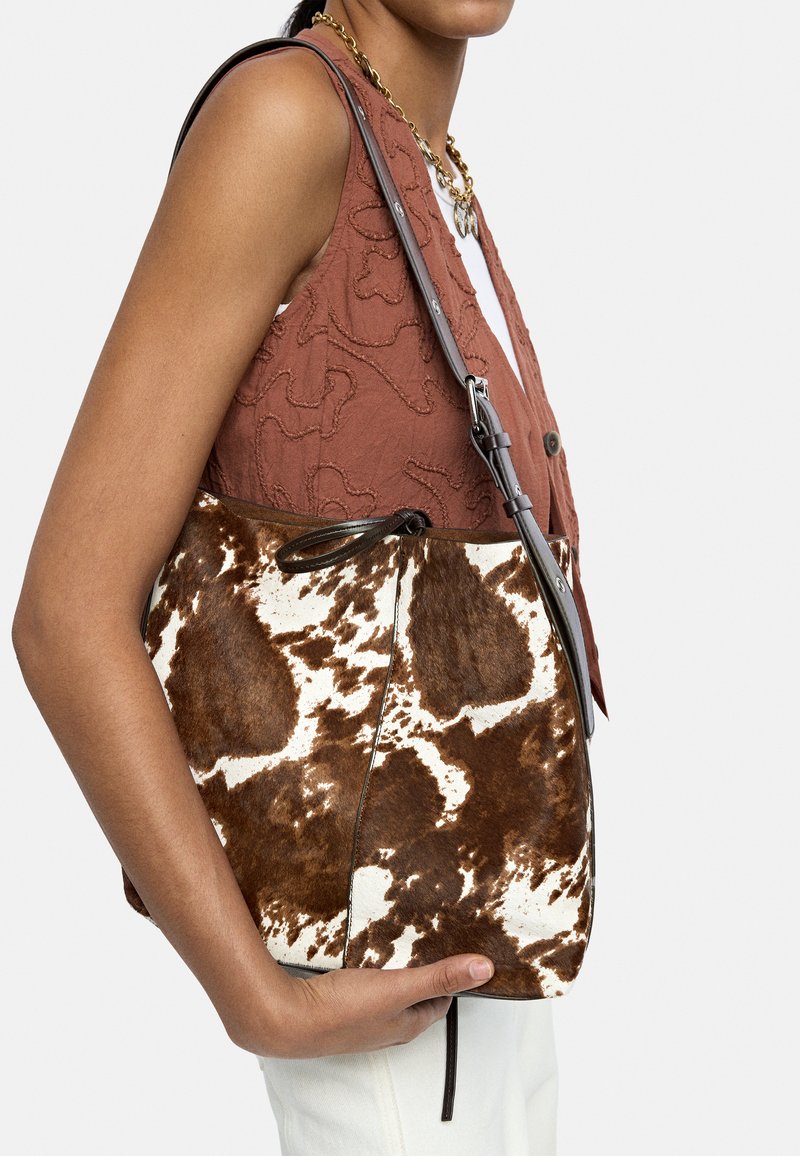 Torba z brązowej i białej skóry bydlęcej o teksturowanym wykończeniu, z pojedynczym paskiem i zapięciem na sznurek, trzymana z boku na tle wzorzystej bluzki.