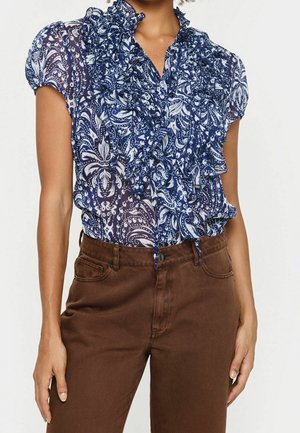 Vrouw draagt een doorschijnende blauw-wit patroon bloes met ruches en bruine broek met hoge taille, staand met handen ontspannen langs haar zij.