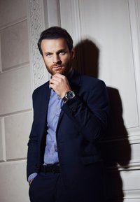 Uomo in un abito scuro con una camicia azzurro chiaro posa, tenendosi il mento. Un orologio metallico con un quadrante blu è in evidenza sul suo polso.