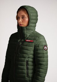 Grøn pufferjakke med høj krave og hætte, med lynlås lukning og to sidelommer. Bemærkelsesværdig rød logo og rundt mærke på ærmet.