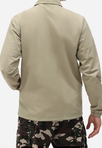 Beige lättviktsjacka med standardkrage och elastiska ärmmuddar, visad bakifrån. Slät textur. Kombinerad med camo cargobyxor.