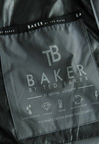Graues Stoffetikett mit der Aufschrift "Baker by Ted Baker". Enthält Symbole für wasserabweisend, langlebig, warm und recyceltes Füllmaterial sowie Pflegehinweise.