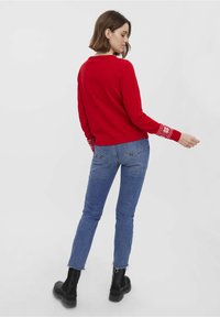 Pull rouge avec des poignets blancs à motifs, porté avec un jean bleu et des bottines noires. Design simple, texture douce, coupe courte.
