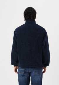 Marineblauer Fleece-Pullover mit hohem Kragen, strukturiertem Stoff und überschnittenen Schultern, kombiniert mit blauen Jeans. Die Rückansicht zeigt eine lockere Passform.