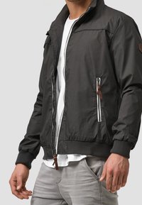 Veste bomber noire avec fermeture éclair sur le devant, poignets et ourlet côtelés. Comprend des poches latérales avec fermetures éclair blanches et tirettes en cuir marron. Tissu texturé.