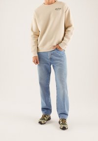 Felpa beige con logo verde, jeans dritti azzurro chiaro e sneaker verdi con motivo. Tessuto morbido e vestibilità comoda.