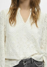 Weiße Spitzenbluse mit V-Ausschnitt, langen Ärmeln, strukturierten geometrischen Mustern und lockerem Schnitt. Kombiniert mit dunklen Jeans.