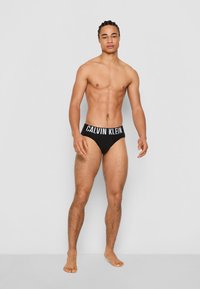 Zwarte katoenen slip met een witte "CALVIN KLEIN" logo tailleband. Model staat rechtop en toont de gladde textuur en het figuurvormende ontwerp.