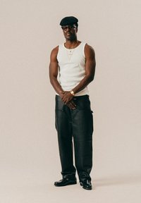 Hvit tanktop, svarte skinnvide bukser, svarte sko, svart flat caps, gullkjeder og solbriller. Enkle farger med minimale design detaljer.