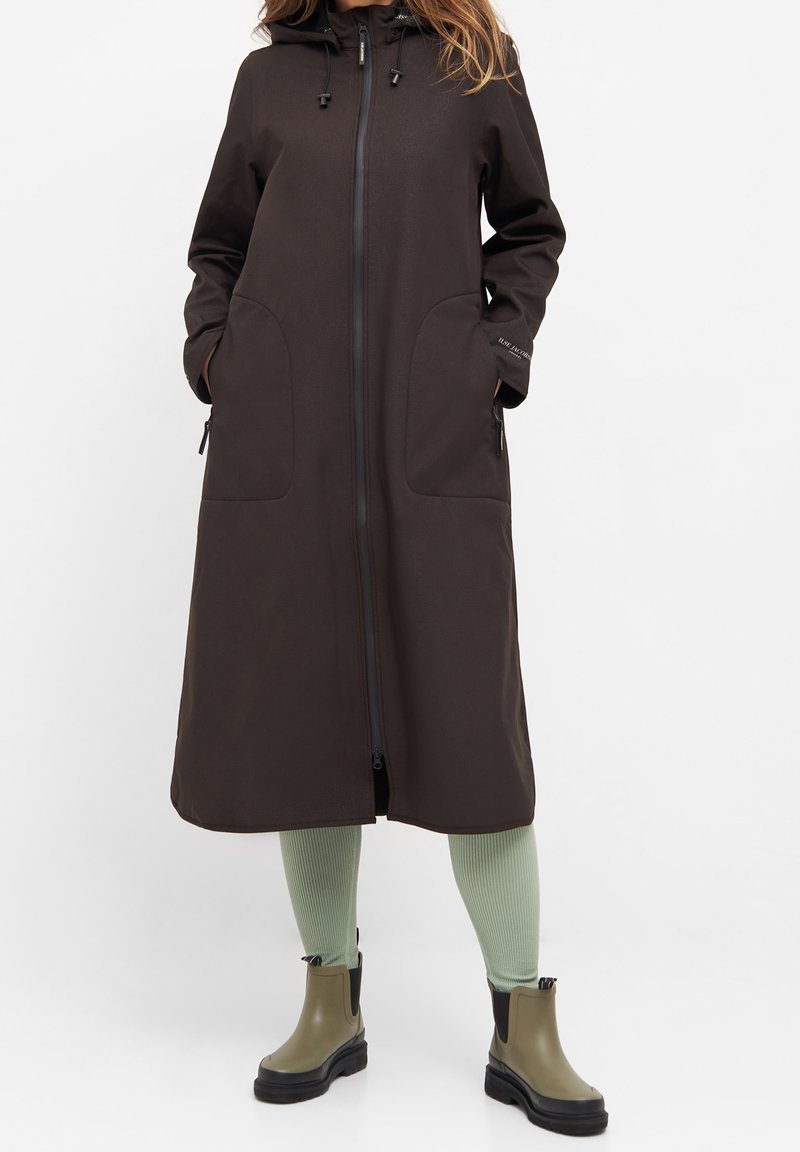 Brauner langer Mantel mit einem Reißverschluss vorne, zwei großen Taschen, einer Kapuze und einer Zug schnur mit Toggle. Getragen über mintgrünen gerippten Leggings und grünen Stiefeln.