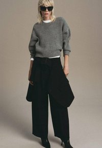Una donna con capelli biondi indossa un maglione grigio oversize, pantaloni neri a gamba larga con particolare tessuto asimmetrico, stivali neri con tacco e occhiali da sole neri.