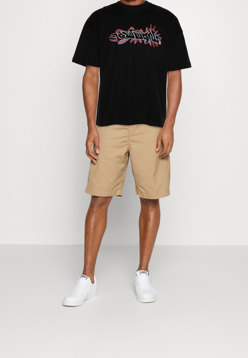 Sort bomulds t-shirt med farverig grafisk tryk, kombineret med beige shorts og hvide sneakers. Afslappet pasform. Glat tekstur.