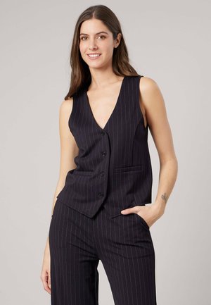 Jonge vrouw met lang bruin haar, gekleed in een zwart mouwloos vest met fijne strepen en bijpassende broek, glimlachend met één hand in de zak.