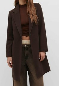 Bruin wollen jas met een getailleerd silhouet, met gesneden revers, een enkele knoopsluiting en lange mouwen, gedragen over een donkere top en broek.