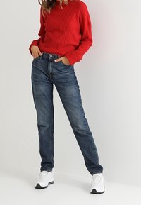 Czerwony prążkowany sweter, niebieskie dżinsy z denimu, białe trampki. Spodnie mają prosty krój nogawek, z efektem sprania i klasycznym wzorem z pięcioma kieszeniami.