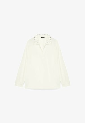 Chemise polo à manches longues blanche avec un col orné de rivets noirs. Fabriquée en tissu doux avec une coupe décontractée, elle possède un plastron avant et des fentes sur les côtés.