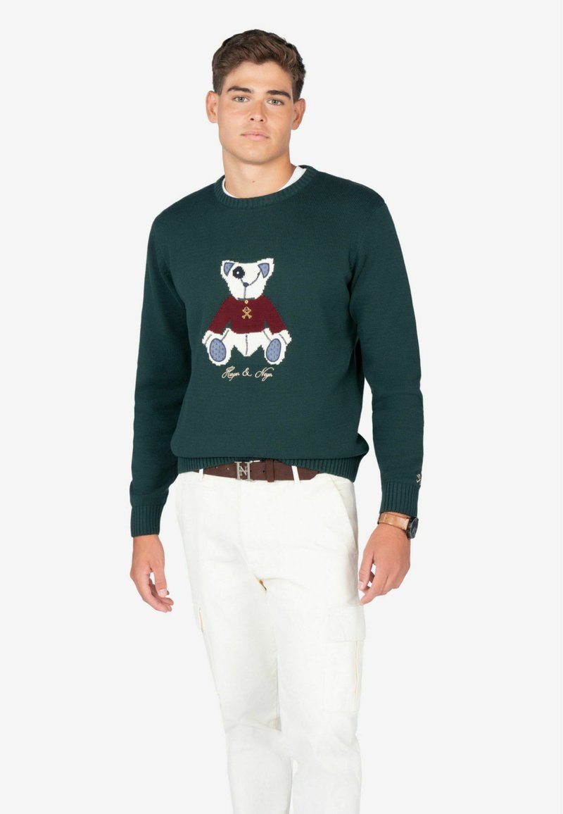 Suéter de punto verde oscuro con un diseño de oso en blanco y burdeos, con texto bordado debajo. Combinado con pantalones cargo blancos.