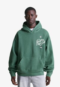 Zelena prevelika kapuca z sprednjim žepom, z grafiko "Srca ne lažejo" in zvezdico na prsih. Bombažni material, mehak na otip.