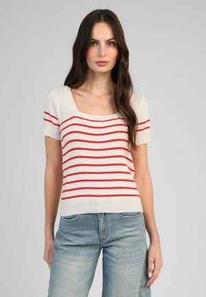 Femme aux longs cheveux bruns portant un haut blanc à manches courtes avec des rayures rouges horizontales et un jean bleu clair, debout devant un fond uni.