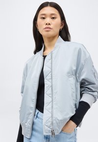 Pitkätummat hiukset omaava nainen, joka käyttää hopeanväristä bomber-takkia mustan paidan ja vaaleansinisten farkkujen päällä, seisoo käsi yhdessä taskussa.