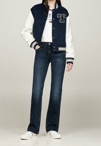 Chaqueta varsity en azul marino y blanco con cuerpo de lana, puños de punto y detalles bordados, llevada sobre un suéter blanco y combinada con jeans de mezclilla oscuros y acampanados.
