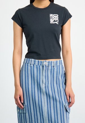 Kobieta ubrana w czarny krótki t-shirt z napisem „OBEY NOTHING” oraz niebiesko-białą spódnicę w pionowe paski z bocznymi kieszeniami.