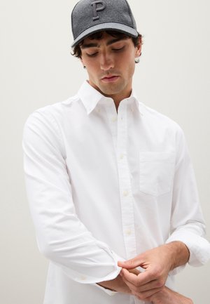 Camisa blanca de botones con mangas largas, un bolsillo en el pecho y textura suave. Gorra gris con una letra "P" elevada en la parte frontal.