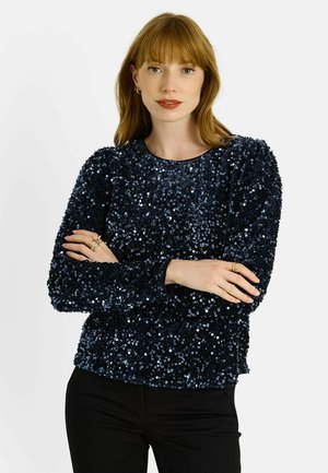 Top de lentejuelas azul marino con escote redondo, mangas largas y superficie texturizada. Presenta una silueta ajustada y pantalones de tela negra.