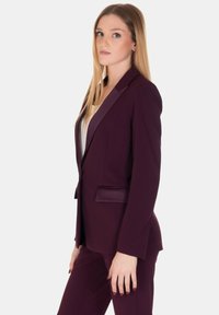Blazer tailored bordeaux con rever a punta, chiusura a un bottone e tasca frontale. Texture liscia, forma aderente.